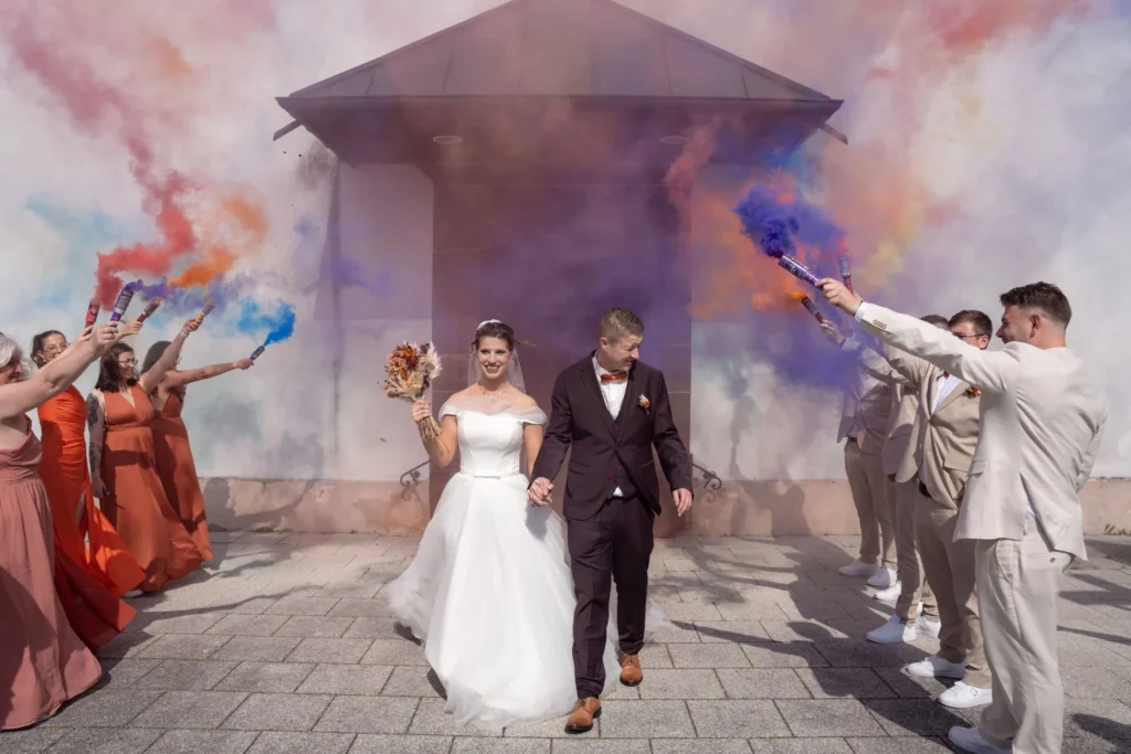 Des mariés sortent de la cérémonie, entourés de leurs proches et de fumigènes colorés